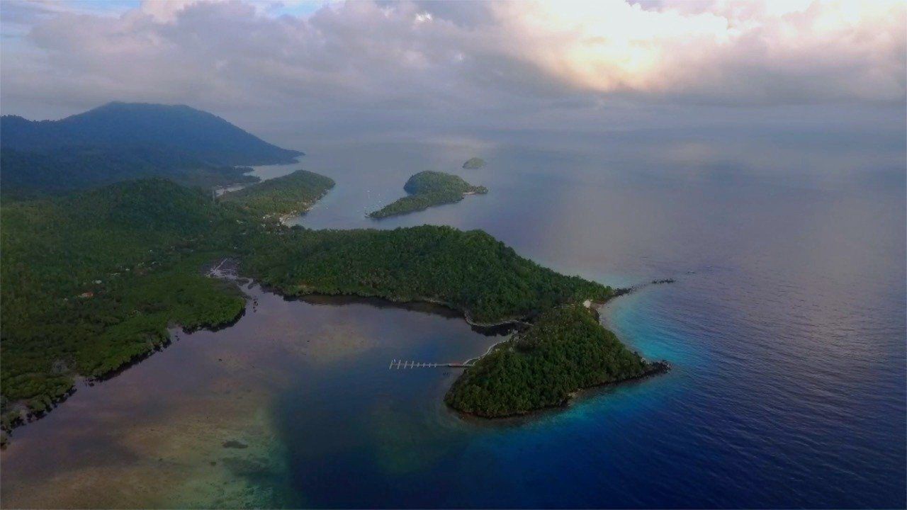 The northwestern coast of Pulau Weh from the air at dusk: forested headlands, a jetty running out over the shallows and the bay turning from turquoise to deep blue.