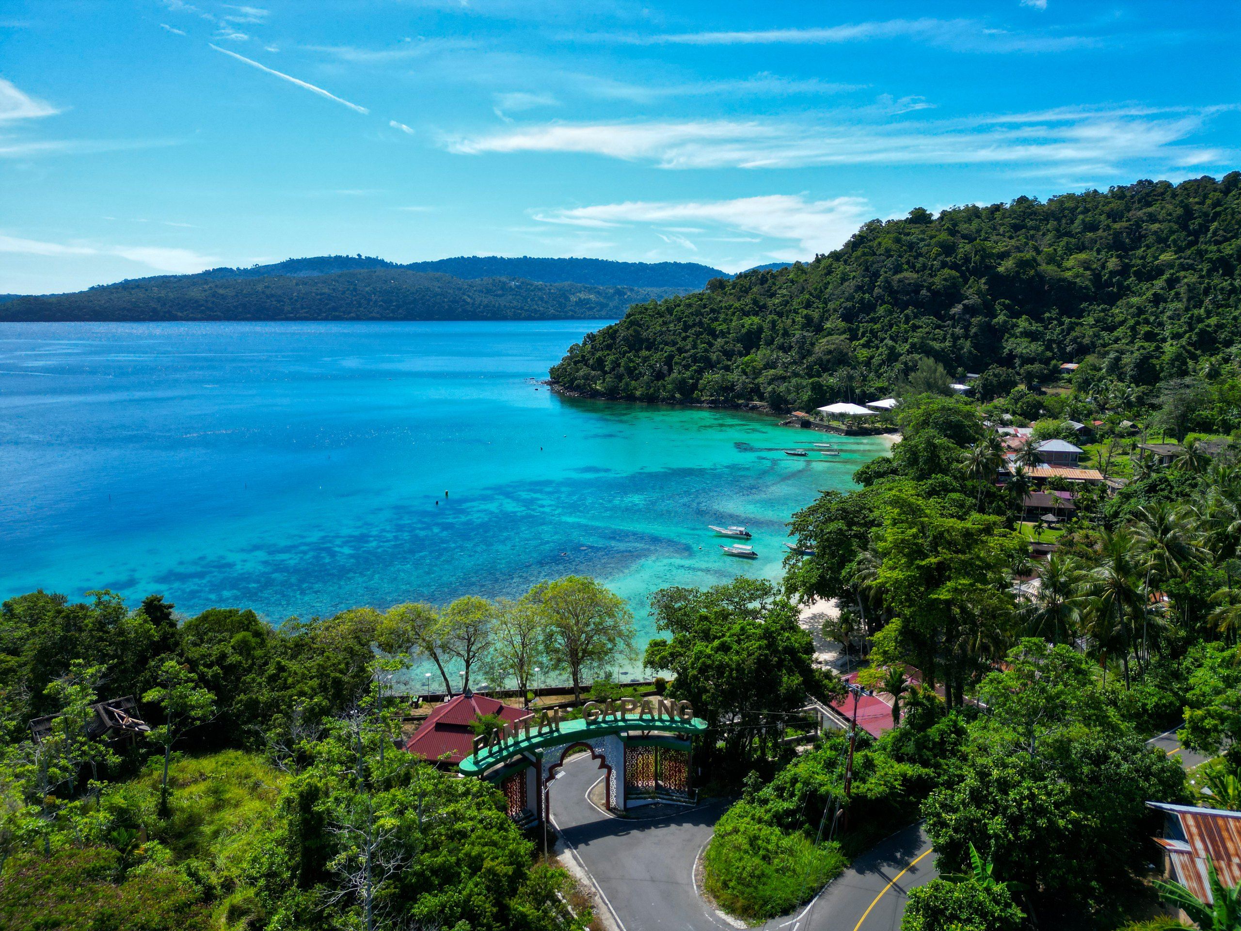 Gapang bay from the air: the Pantai Gapang gate on the coast road, boats moored over turquoise shallows, forested hills behind.