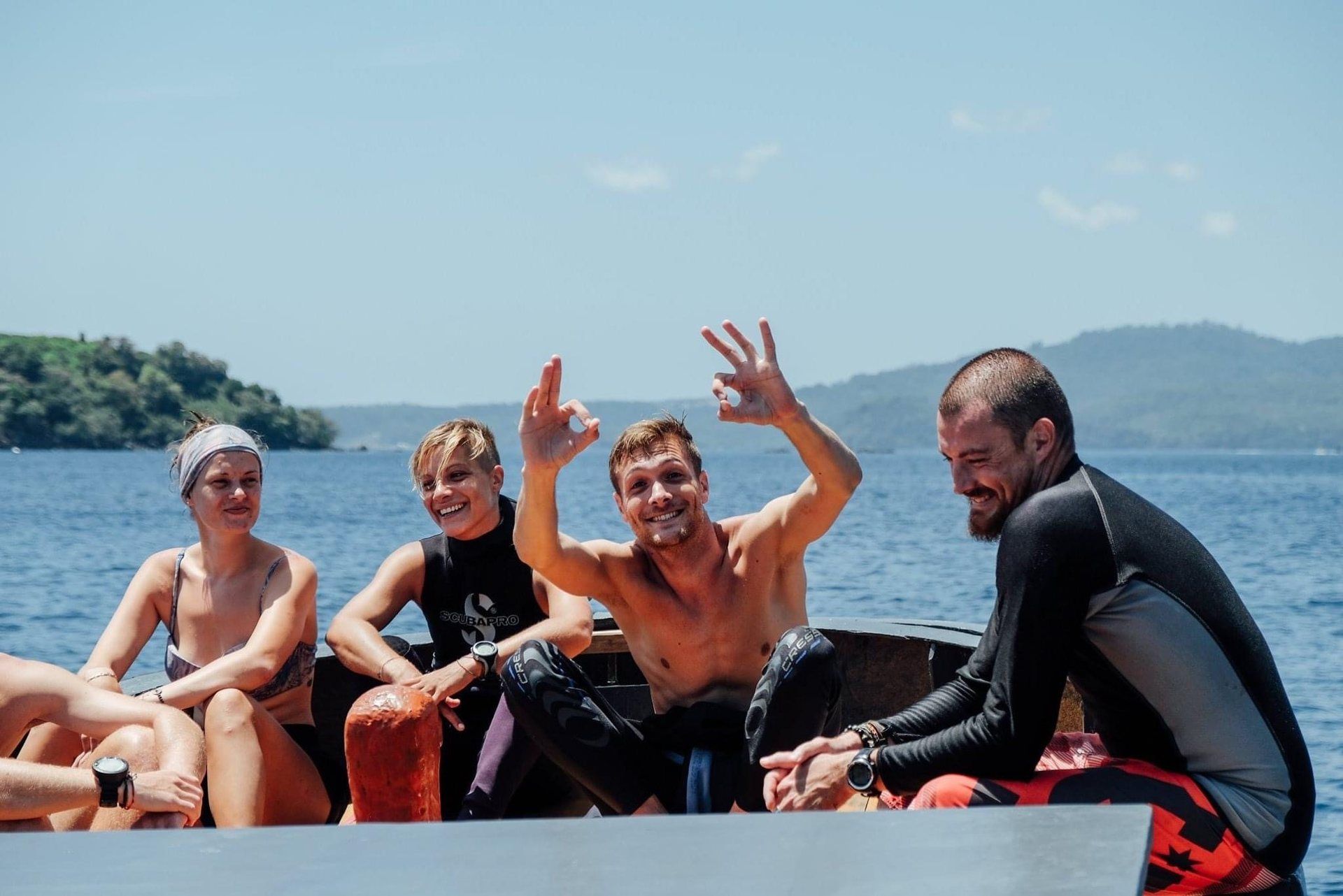 Four divers sitting on the dive boat after a dive, one of them giving the OK sign, with the coast of Pulau Weh behind them.