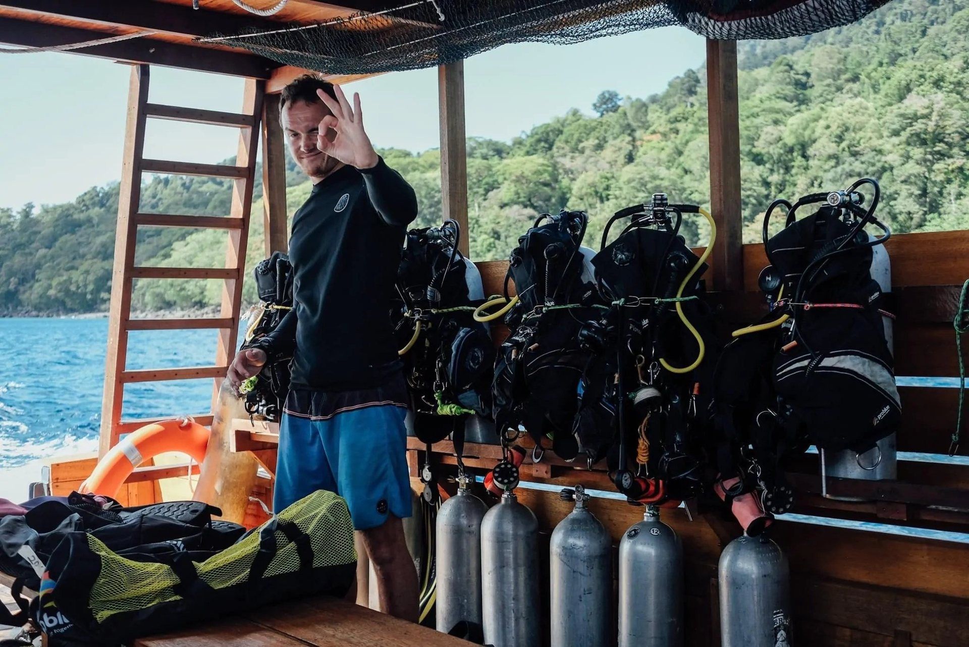Assembled scuba sets and tanks racked along the gear bench on the boat, a crew member signalling OK