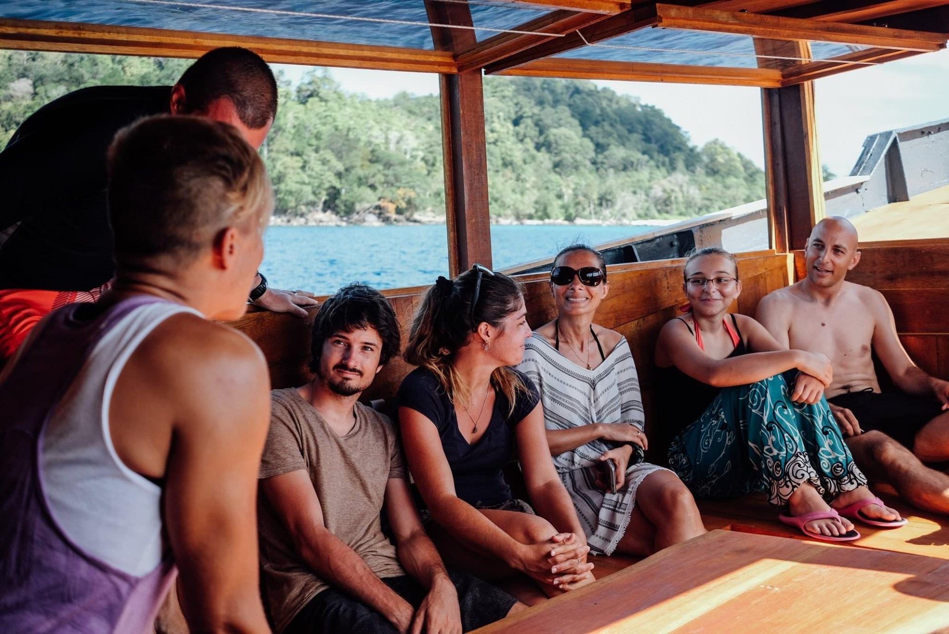Guests sitting together in the shaded cabin of the dive boat, some in dive gear and some not, with the forested coast of Pulau Weh through the open side.