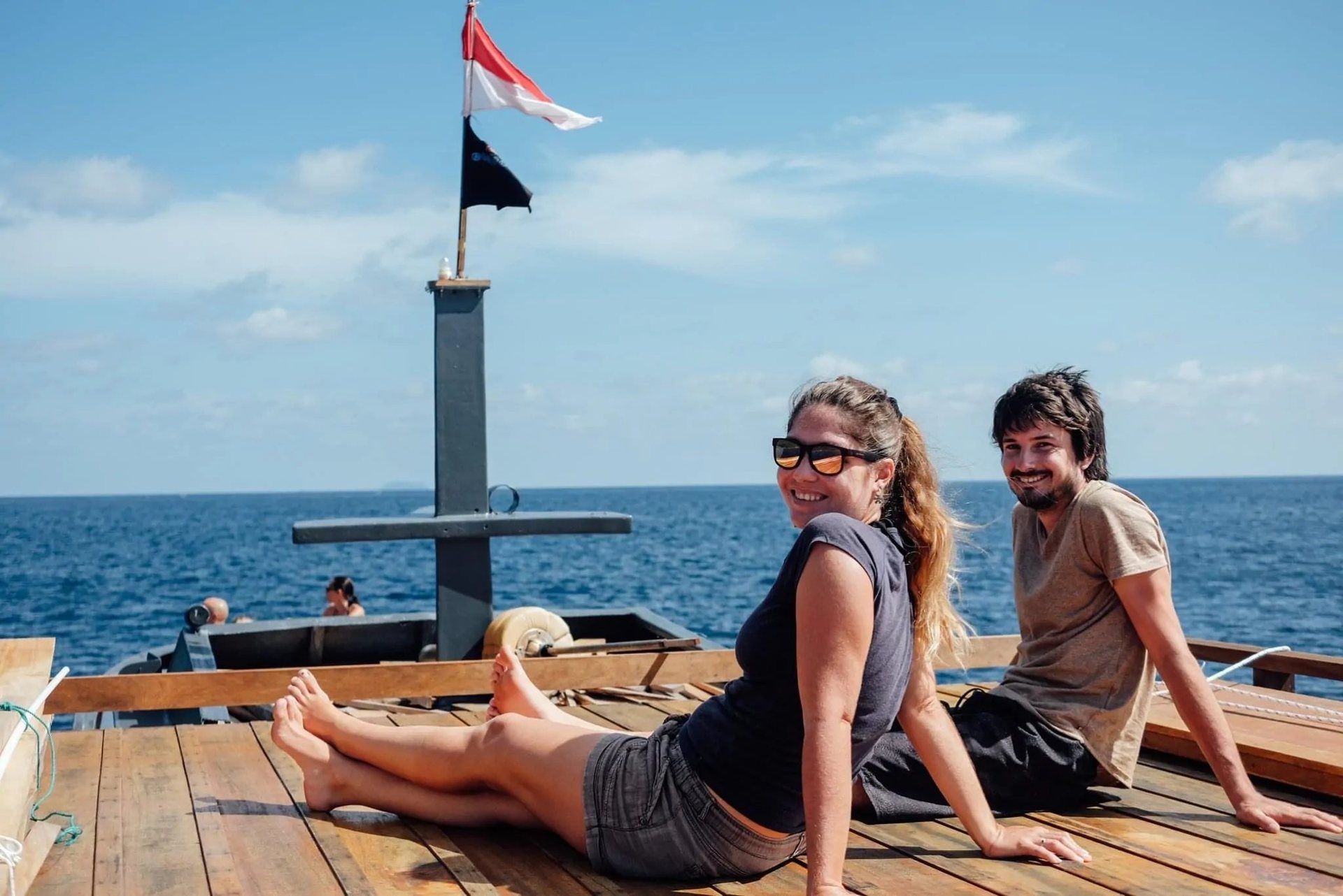 Two divers sitting on the wooden rooftop deck of the dive boat in open water, the Indonesian flag flying from the mast behind them.