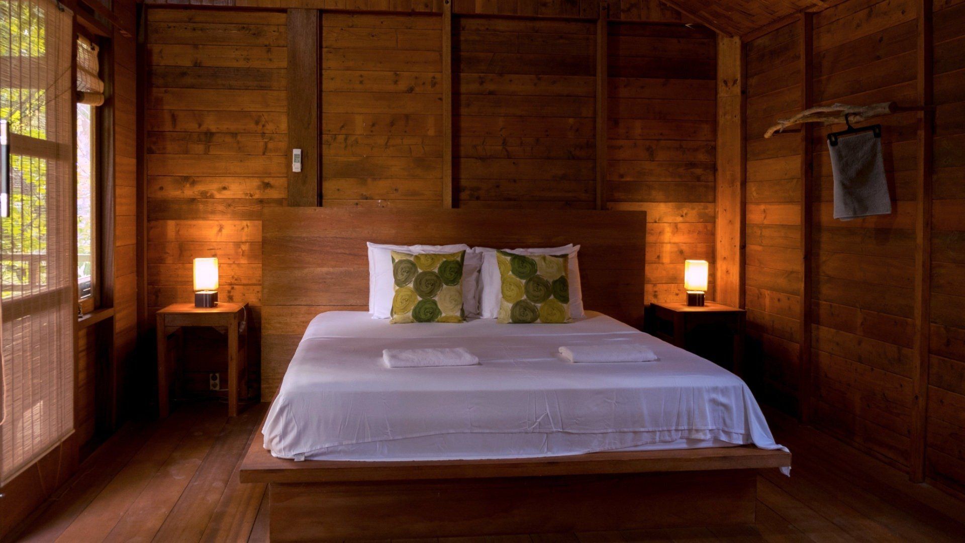 Inside one of the wooden bungalows, a double bed made up with white linen between two lit bedside lamps.