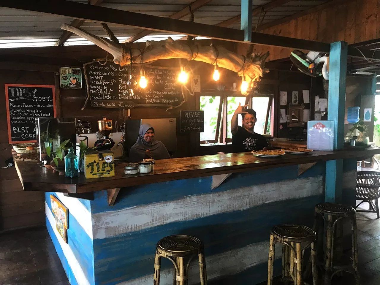 The café bar at the dive centre: a driftwood beam strung with bulbs over the counter, the drinks menu chalked on the board behind it, two staff at work.