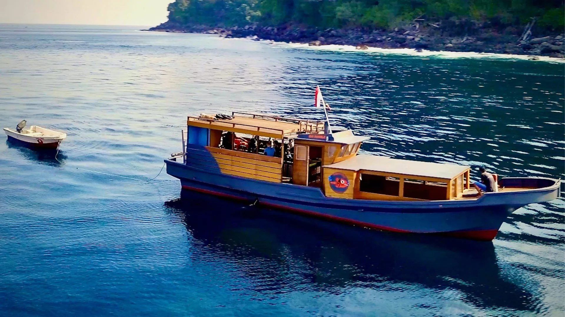 A wooden dive boat with a rooftop deck, anchored off a rocky shore.