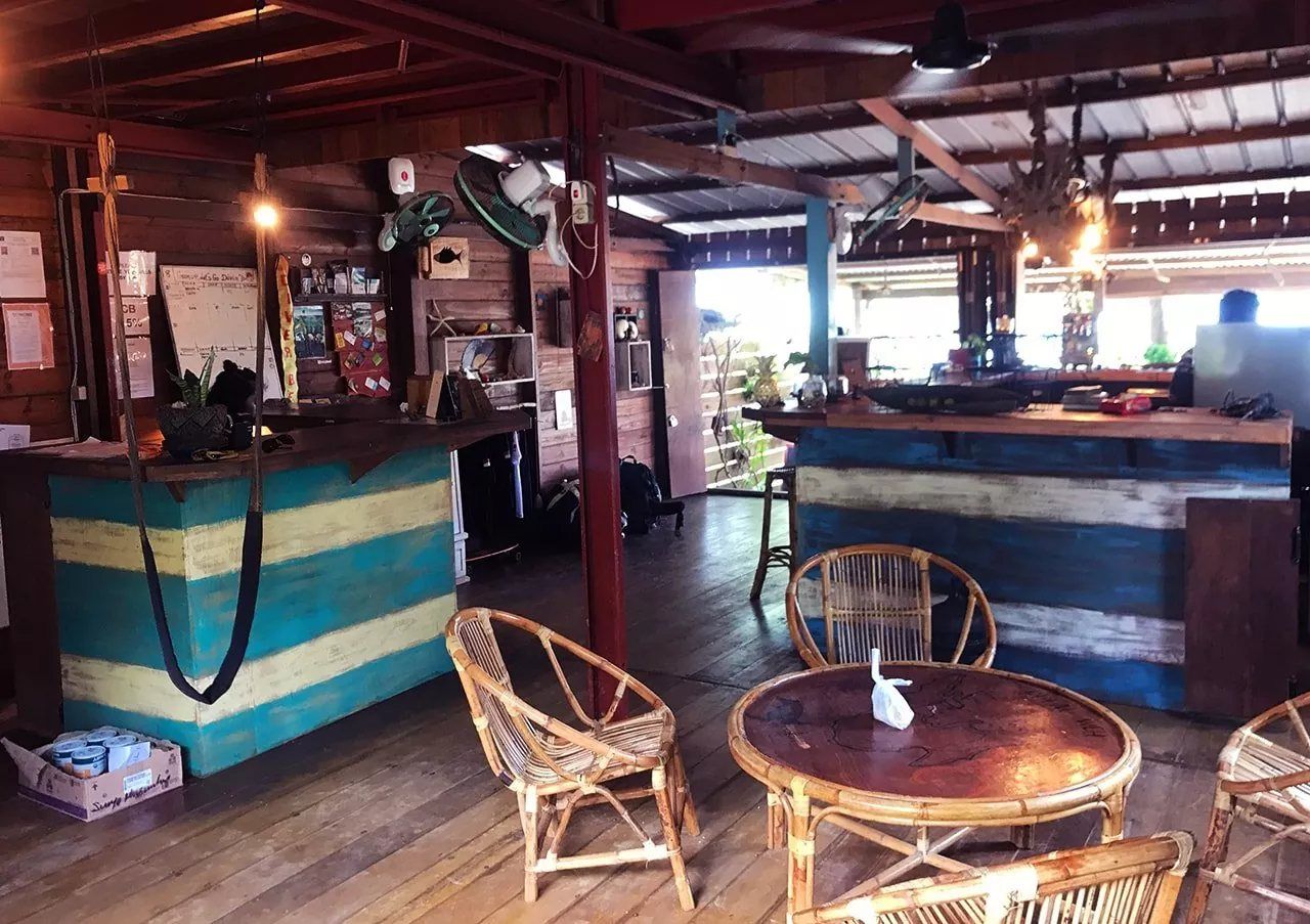 The reception and lounge at the dive centre: a timber counter painted in bands of blue and cream, rattan chairs around a low table and fans under the roof beams.