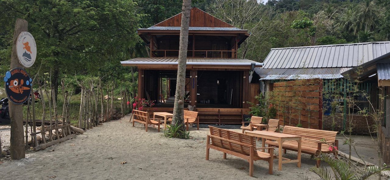 The Bubble Addict dive centre at Gapang Beach: the timber building open to the sand, teak benches out front and the round dive centre sign on its post.